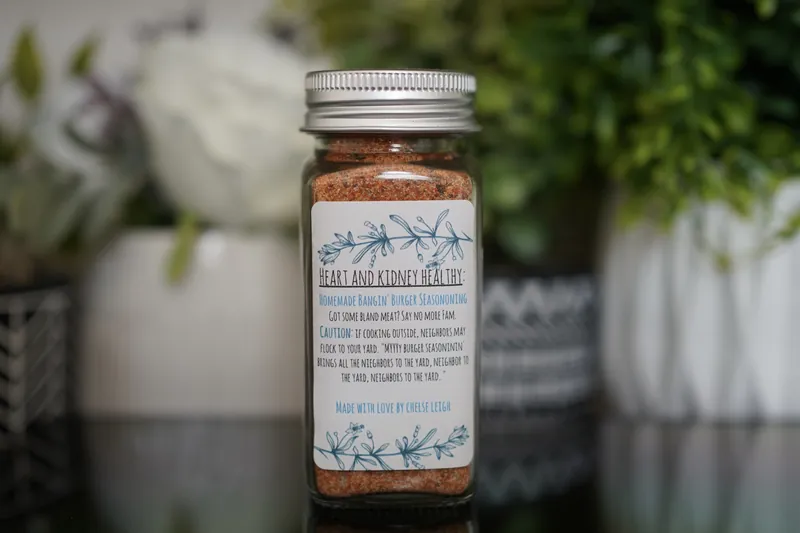 A glass jar of Homemade Bangin' Burger Seasoning, warm red-orange, with a silver lid and a white botanical label, on a dark reflective counter with greenery behind.