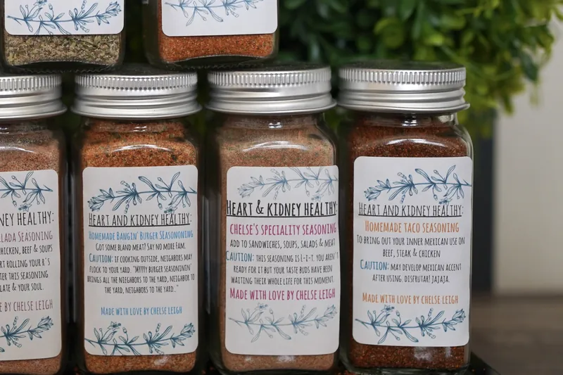 A glass jar of Chelse's Speciality Seasoning among the other blends, its pink-lettered white label facing forward on a dark reflective counter.