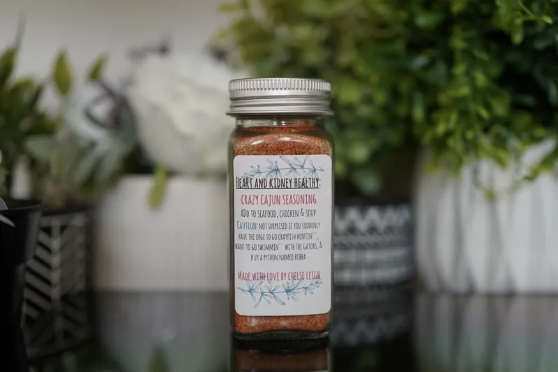 A glass jar of Crazy Cajun Seasoning, deep orange-red, with a silver lid and a white botanical label, on a dark reflective counter with greenery behind.