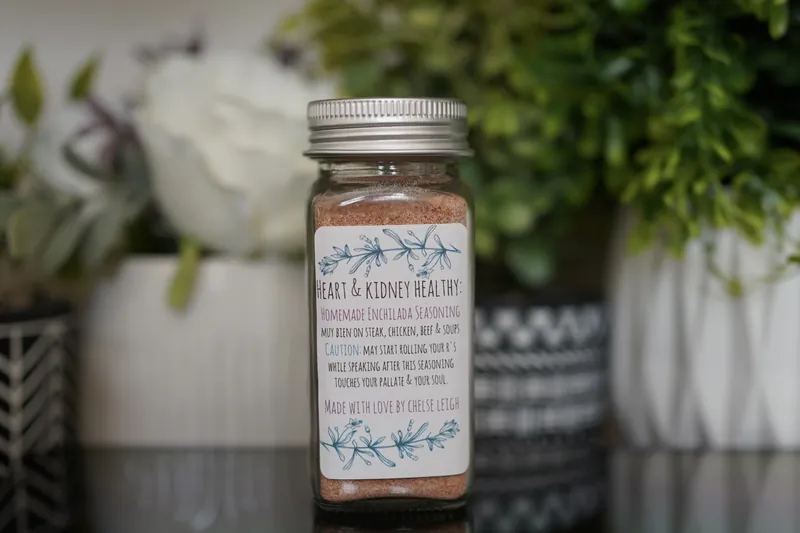 A glass jar of Homemade Enchilada Seasoning, deep red, with a silver lid and a white botanical label, on a dark reflective counter with greenery behind.