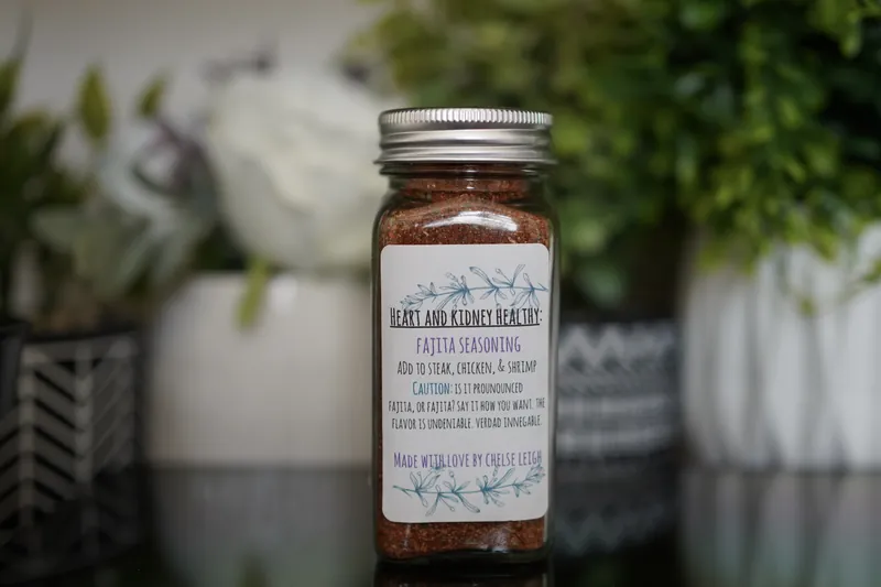 A glass jar of Fajita Seasoning, bright red-orange, with a silver lid and a white botanical label, on a dark reflective counter with greenery behind.