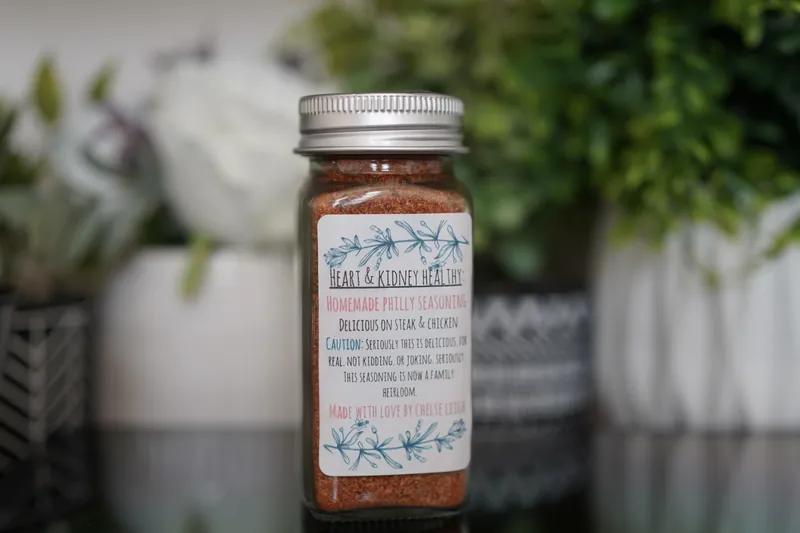 A glass jar of Homemade Philly Seasoning, rust-coloured, with a silver lid and a white botanical label, on a dark reflective counter with greenery behind.