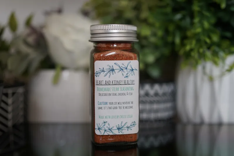 A glass jar of Homemade Steak Seasoning, deep red-brown, with a silver lid and a white botanical label, on a dark reflective counter with greenery behind.