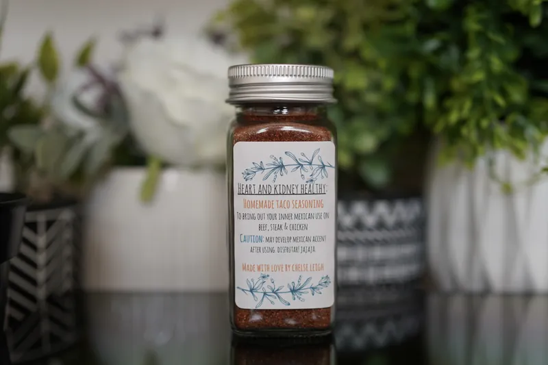 A glass jar of Homemade Taco Seasoning, brick red, with a silver lid and a white botanical label, on a dark reflective counter with greenery behind.