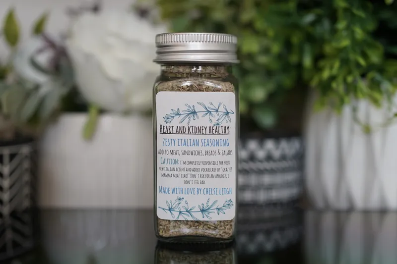 A glass jar of Zesty Italian Seasoning, flecked green and gold, with a silver lid and a white botanical label, on a dark reflective counter with greenery behind.